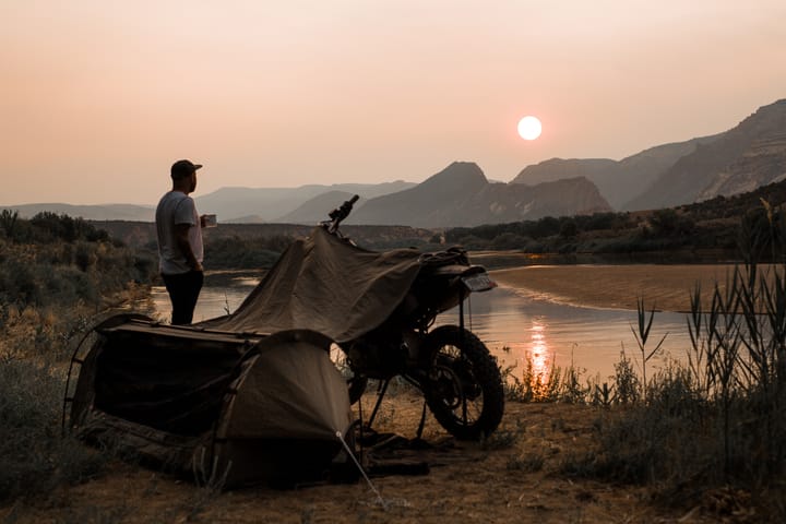 a man watches the sunset next to a motorcycle and a tent