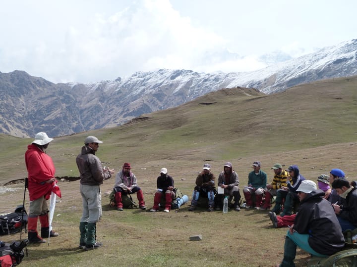 a group of outdoor enthusiasts sitting in a circle in an alpine meadow, listening to two instructors