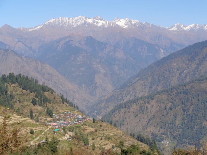 a hillside village in a forested valley, with snow-capped peaks in the backdrop