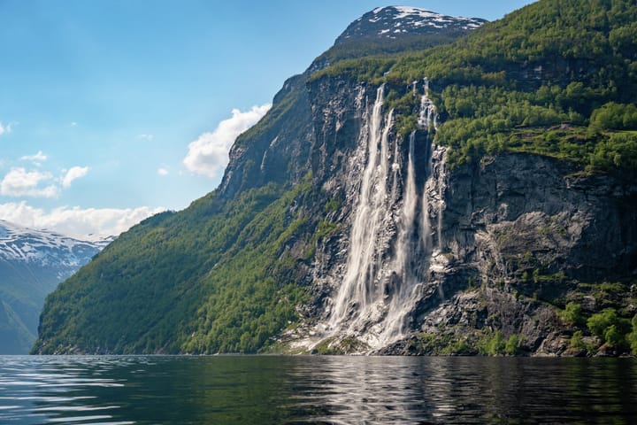 a waterfall streams down a forested cliff into a massive fjord